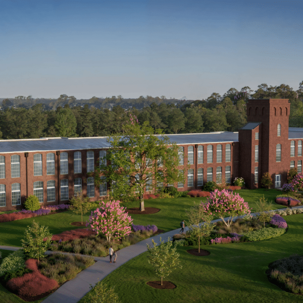 A red-brick building with tall windows and a central tower stands in lush gardens. Cars park nearby, trees and blue sky behind.