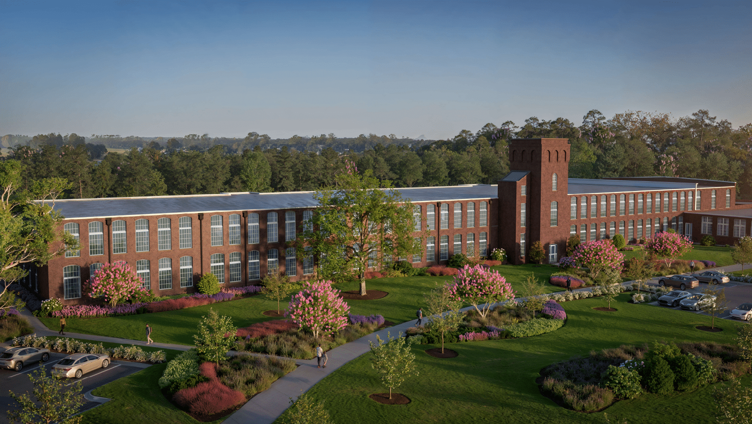 A red-brick building with tall windows and a central tower stands in lush gardens. Cars park nearby, trees and blue sky behind.