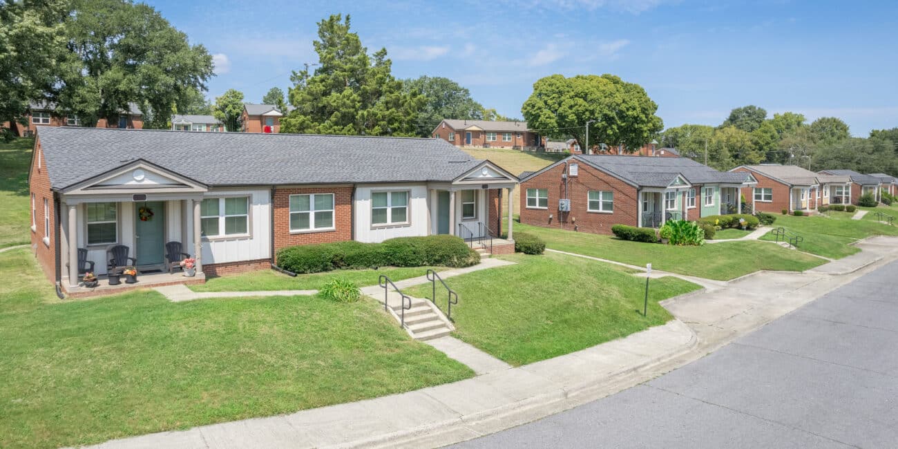 Single-story brick houses with neat lawns line a quiet, curved street under a blue sky; steps lead to porches, trees in back.