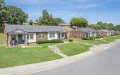 Single-story brick houses with neat lawns line a quiet, curved street under a blue sky; steps lead to porches, trees in back.