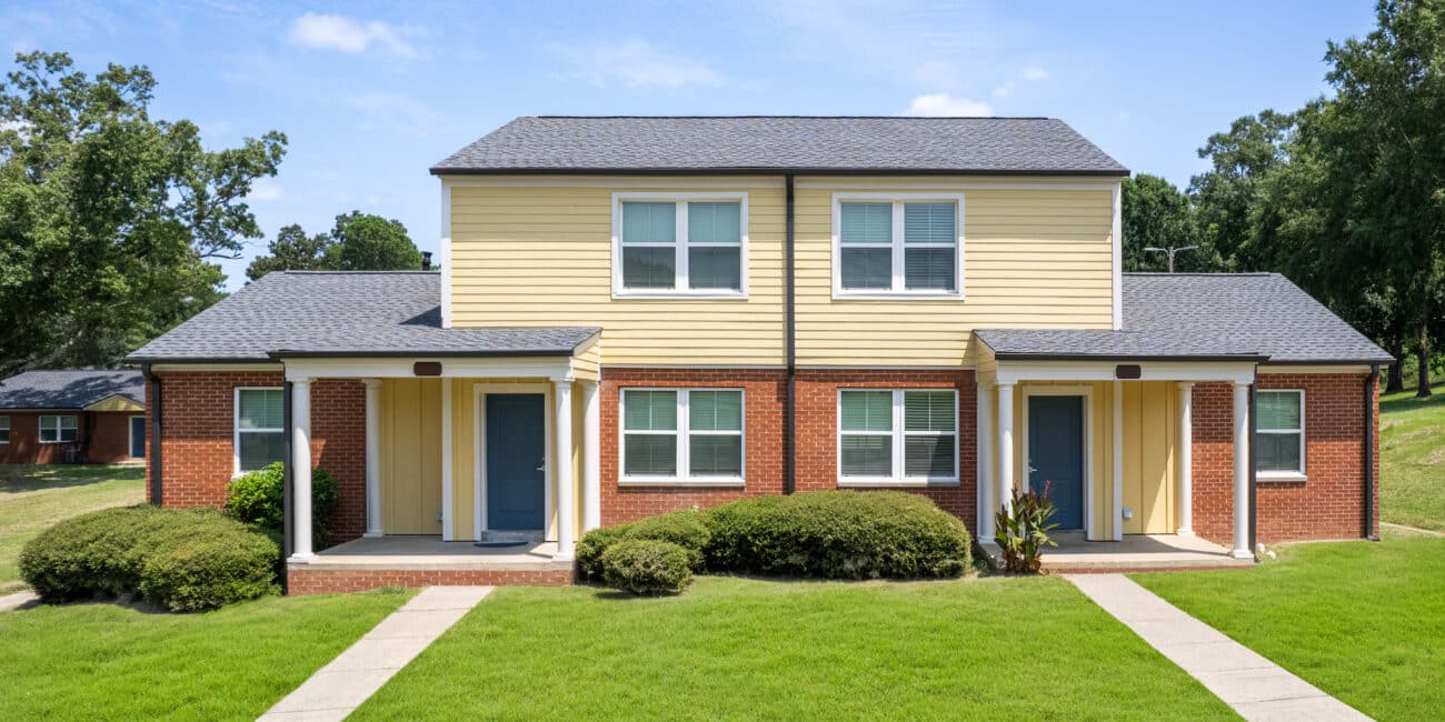 A duplex with red brick and yellow siding, twin front doors, white trim, small porches, green grass, shrubs, trees beneath blue sky.