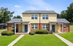 A duplex with red brick and yellow siding, twin front doors, white trim, small porches, green grass, shrubs, trees beneath blue sky.
