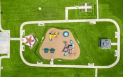 Aerial view of a green park with central playground, walking paths, benches, gazebo, star-shaped feature, and flowerbeds on lawn.
