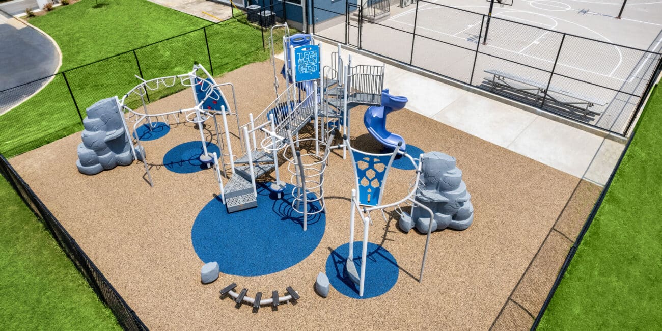 Aerial view of a modern playground with slides, climbing structures, and rubber floor by a basketball court and blue building on grass.