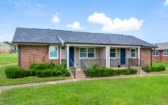 A brick duplex with two doors, small porches, black railings, and front bushes sits on a grassy lawn under a partly cloudy blue sky.