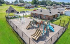A fenced playground with slides, climbing structures, and swings on gravel near a brick building; pavilion and grassy area nearby.