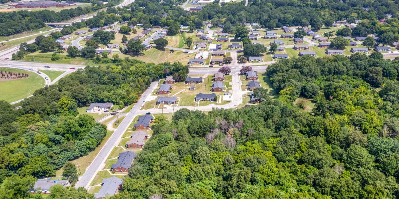 Aerial view of a neighborhood with houses, roads, driveways, and lush green trees under a clear blue sky.