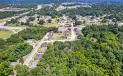 Aerial view of a neighborhood with houses, roads, driveways, and lush green trees under a clear blue sky.