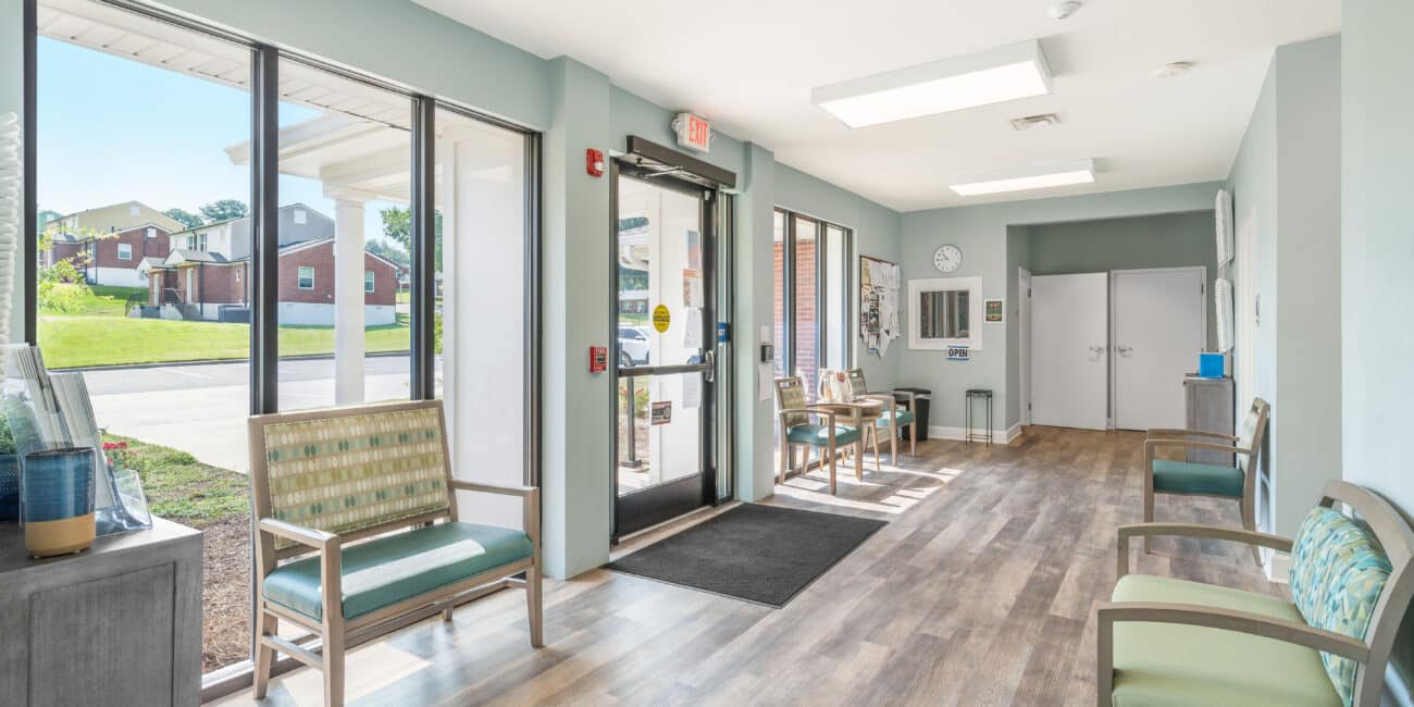 Bright waiting room with large windows, chairs along the walls, wood-pattern floors, brochures, and sunlight creating a welcoming space.