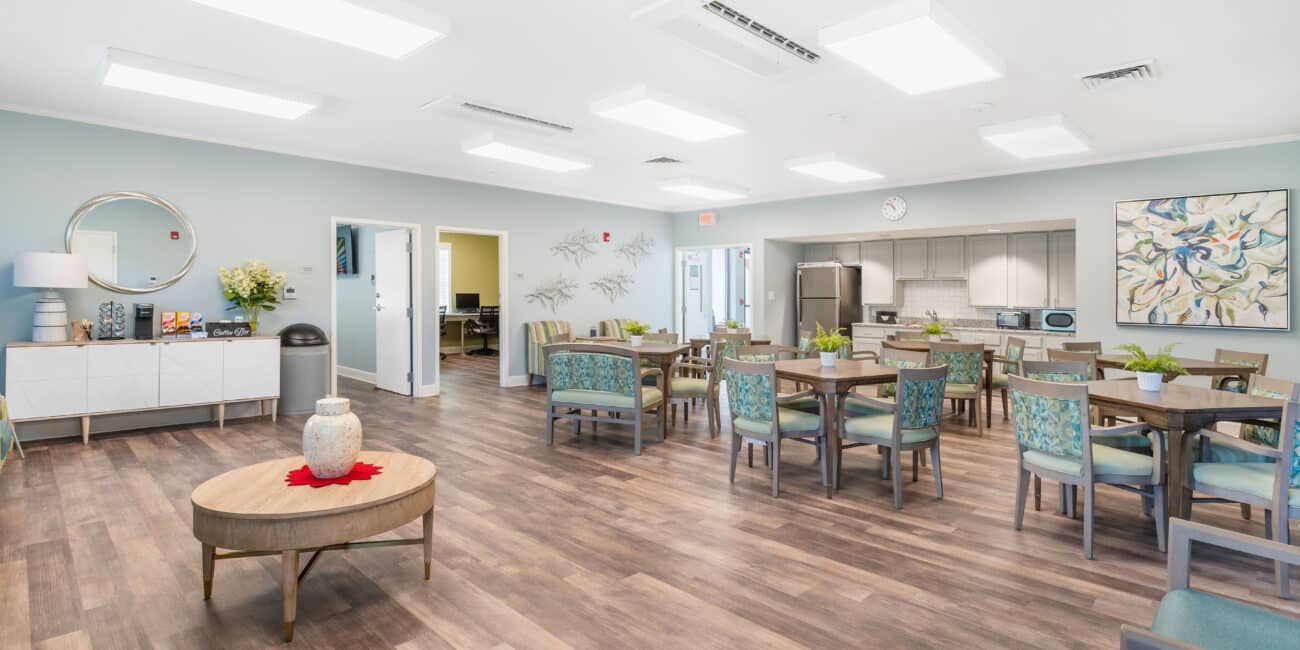 Bright, spacious dining room with wood floors, tables, modern lighting, blue walls, plants, kitchen in back, and wall art.