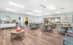 Bright, spacious dining room with wood floors, tables, modern lighting, blue walls, plants, kitchen in back, and wall art.
