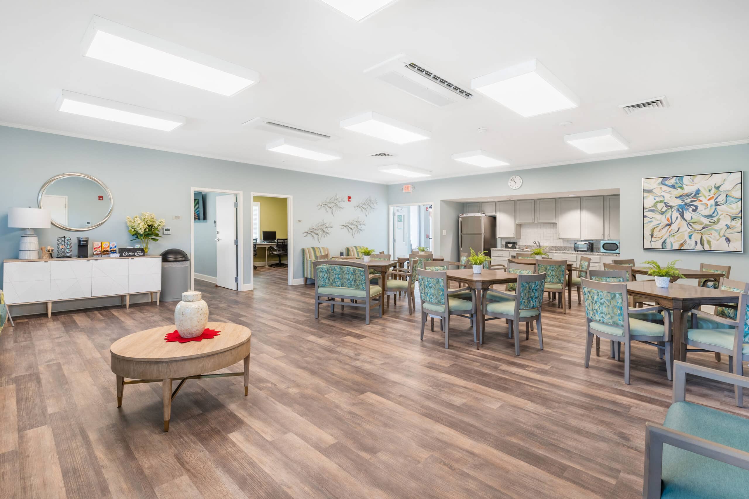 Bright, spacious dining room with wood floors, tables, modern lighting, blue walls, plants, kitchen in back, and wall art.