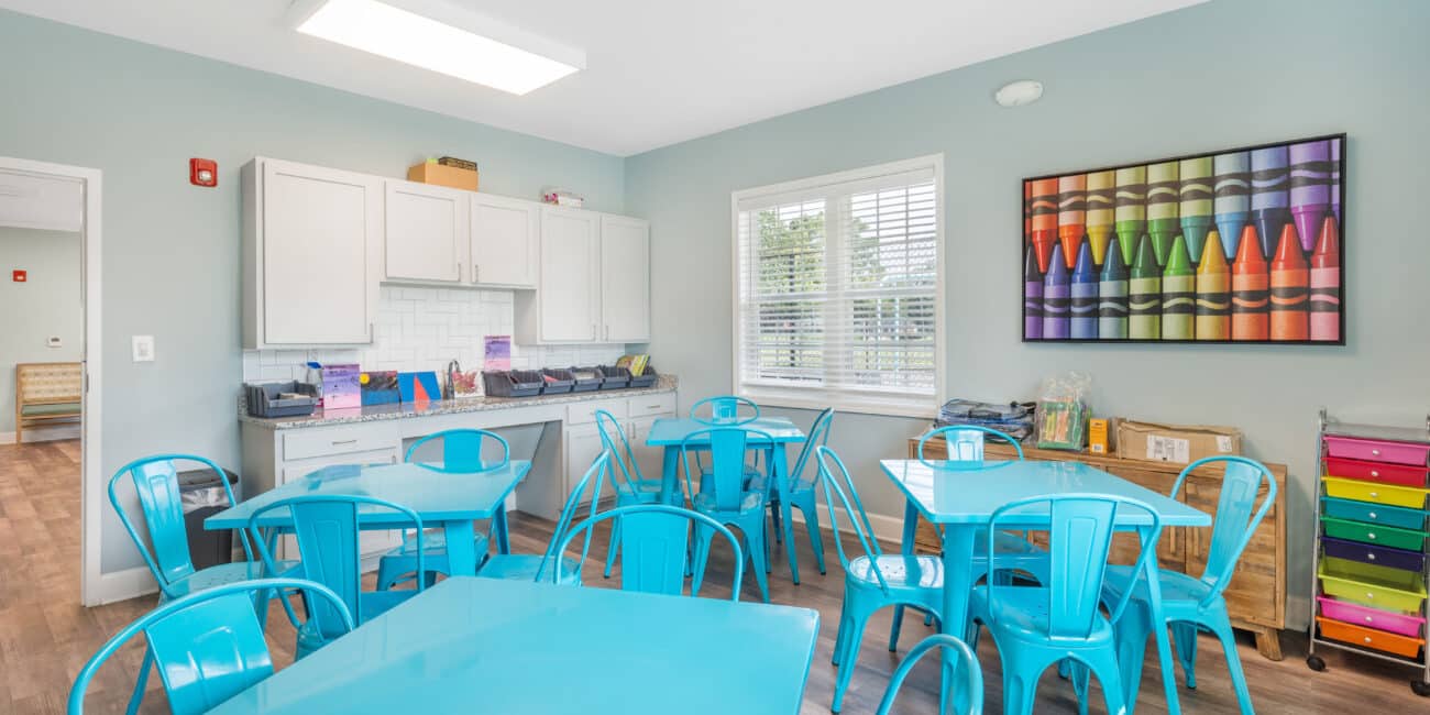 A bright classroom with blue walls, turquoise tables and chairs, storage cabinets, colorful bins, and crayon art. Sunlight streams in.